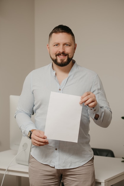 Un homme souriant avec une barbe se tient avec une feuille de papier blanc vide près d'un ordinateur de bureau en acier à la maison