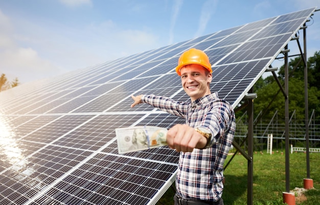 Un homme souriant avec un billet de cent dollars à la main montre à la station de batteries solaires dans lesquelles les rayons du soleil se reflètent