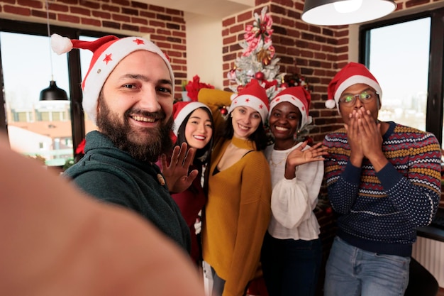 Photo homme souriant en bonnet de noel prenant du slefie avec des collègues dans un bureau décoré lors d'une fête d'entreprise de noël. des collègues regardent la caméra tout en posant pour une photo de groupe sur téléphone portable sur un lieu de travail festif