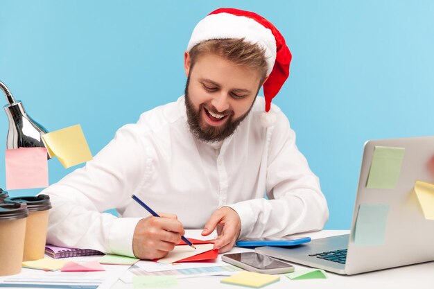 Homme souriant en chapeau de père noël écrivant une lettre ou signant une carte postale de voeux dans une enveloppe rouge, façon romantique de féliciter avec les vacances. Tourné en studio intérieur isolé sur fond bleu