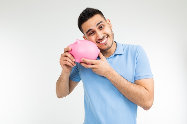 Homme souriant dans un t-shirt bleu embrasse une tirelire sur fond blanc avec copie espace