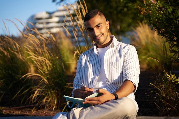 Homme souriant du Moyen-Orient regardant la caméra et prenant des notes sur le cahier assis dans le parc