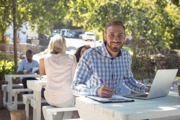 Homme souriant, écrit sur le presse-papiers tout en utilisant un ordinateur portable