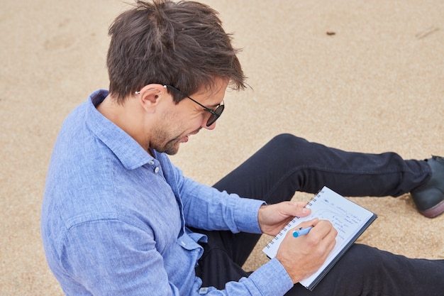 Homme souriant écrivant dans un cahier avec son stylo alors qu'il était assis sur le sable.