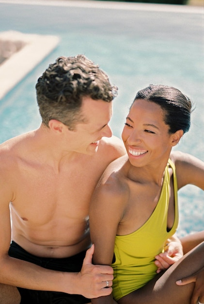 Un homme souriant embrasse une femme heureuse par la taille en la regardant alors qu'il est assis au bord de la piscine.