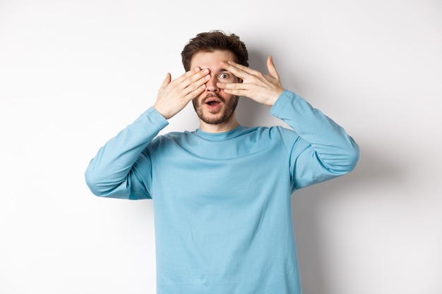 Homme souriant étonné couvrant les yeux avec les mains mais regardant à travers les doigts quelque chose de génial, debout sur fond blanc.