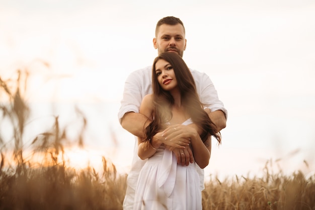 Homme souriant étreignant sa jolie femme tout en se tenant derrière elle sur un champ de blé pendant le coucher du soleil du soir. Concept d'amour