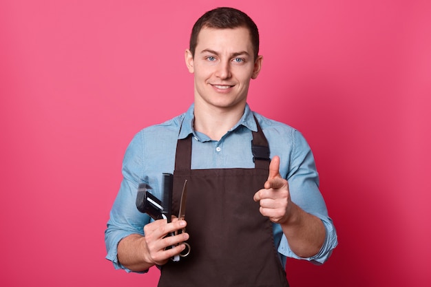 Homme souriant avec une expression satisfaite, vêtu d'un tablier et d'une chemise, détient l'équipement nécessaire pour le coiffeur