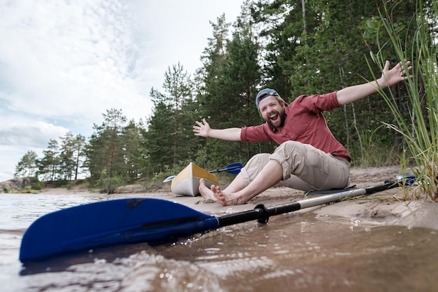 Un homme souriant joyeux se repose sur la rive du lac après une excursion en kayak, il a écarté les bras