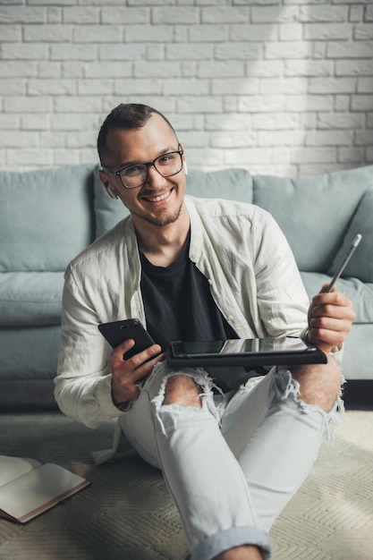 Homme souriant à lunettes travaillant à distance avec tablette numérique et téléphone portable tout en étant assis sur flo