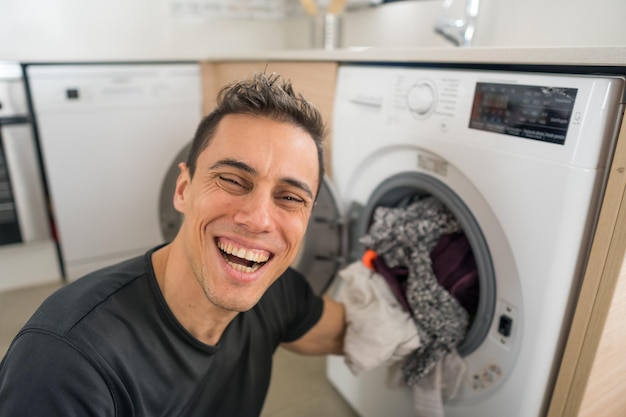 Homme souriant mettant des vêtements dans la machine à laver, dans la cuisine. Fermer.