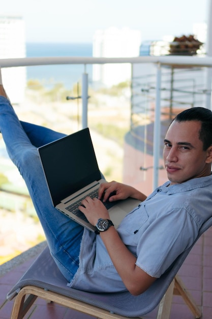 Homme souriant avec ordinateur portable travaillant sur son balcon à la maison