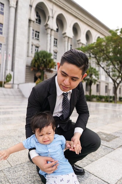 Un homme souriant portant un costume tenant son fils alors qu'il s'accroupit à l'extérieur