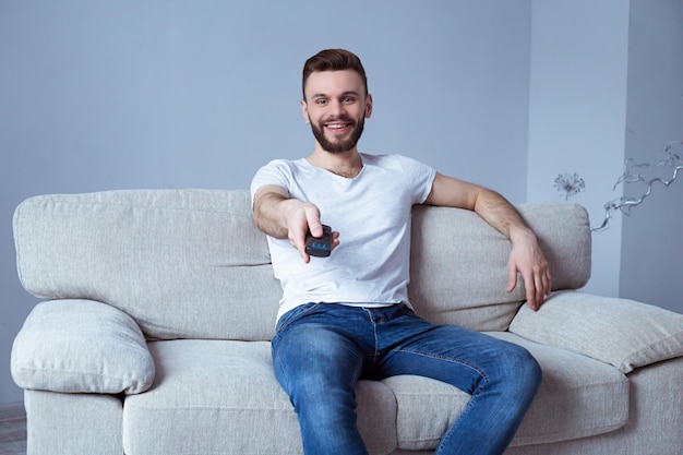 Homme souriant regardant la télévision assis à la maison sur un canapé et changeant de chaîne par télécommande