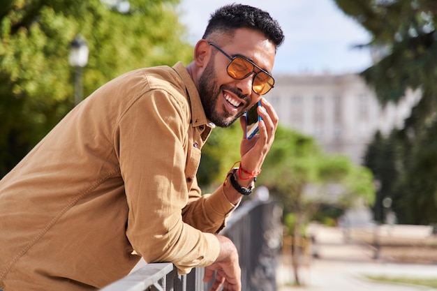 Homme souriant s'appuyant sur une balustrade parlant sur son téléphone portable jeune homme latino avec lunettes de soleil et barbe regardant la caméra