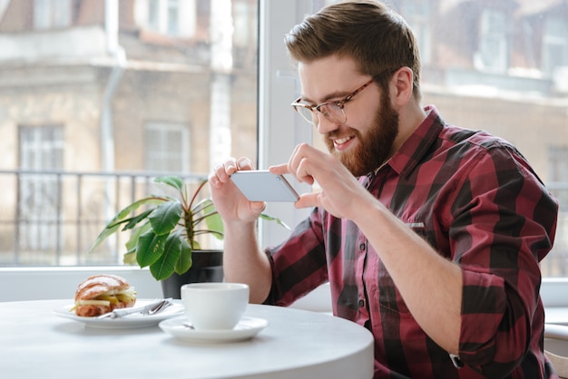 Homme souriant avec smartphone photographiant son déjeuner au café