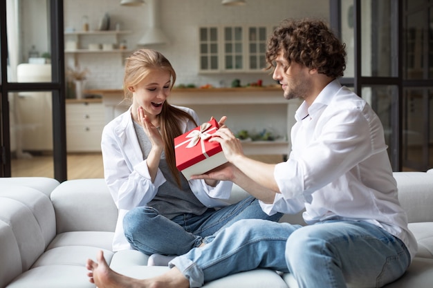 Un homme souriant surprend sa petite amie avec un cadeau à la maison. Donner des cadeaux.