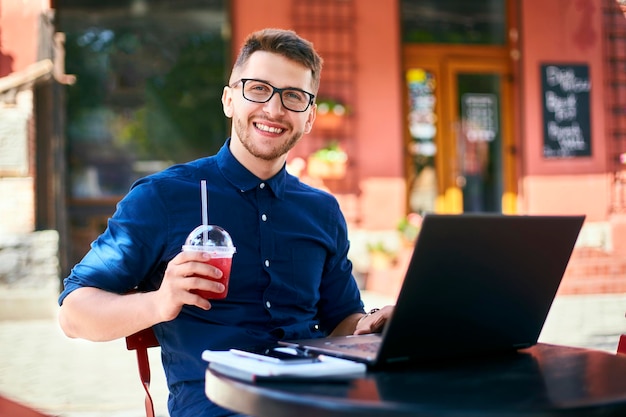 Un homme souriant avec une tasse en plastique de jus de fruits frais à la main travaille avec un homme d'affaires d'ordinateur portable dans des verres à boissons