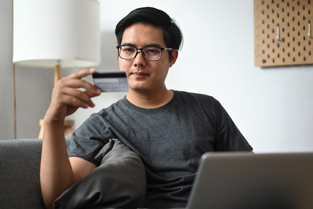 Homme souriant tenant une carte de crédit et faisant des achats en ligne avec un ordinateur portable à la maison.