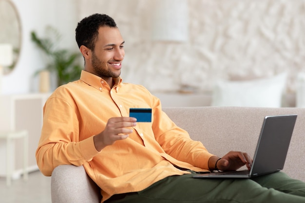 Homme souriant utilisant un ordinateur et une carte de crédit assis sur un canapé