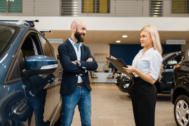 Homme souriant et vendeuse chez le concessionnaire automobile. Client et vendeur dans la salle d'exposition de véhicules, homme achetant des transports, entreprise de concessionnaire automobile