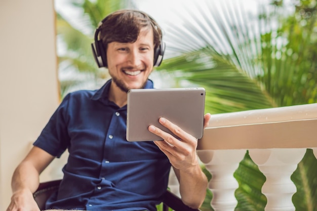 Homme sous les tropiques parlant avec ses amis et sa famille lors d'un appel vidéo à l'aide d'une tablette et d'un casque sans fil assis sur la terrasse.