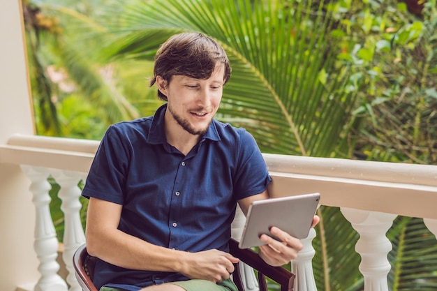 Homme sous les tropiques parlant avec ses amis et sa famille lors d'un appel vidéo à l'aide d'une tablette et d'un casque sans fil assis sur la terrasse.