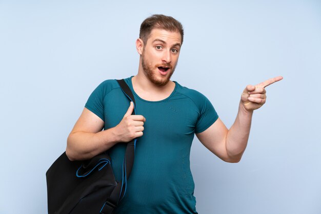 Homme de sport blonde sur le mur bleu surpris et pointant le doigt vers le côté