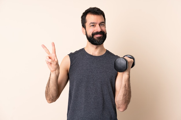 Homme de sport caucasien avec barbe faisant de l'haltérophilie sur un espace isolé montrant le signe de la victoire avec les deux mains