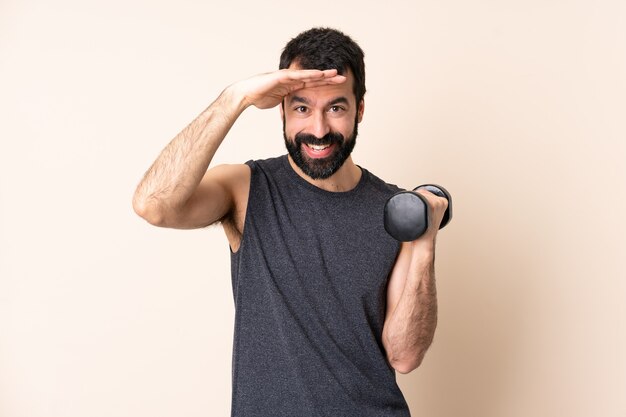 Homme de sport caucasien avec barbe faisant de l'haltérophilie sur un mur isolé à la recherche de loin avec la main pour regarder quelque chose