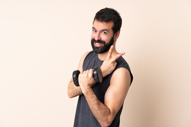 Homme de sport caucasien avec barbe faisant de l'haltérophilie sur le mur en souriant et montrant le signe de la victoire