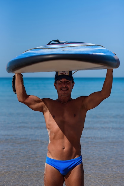 Homme sportif bronzé tient sa planche de surf au dessus de sa tête sur la mer.