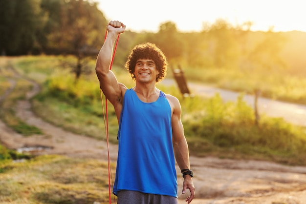 Homme sportif caucasien souriant aux cheveux bouclés, faire des exercices d'étirement avec une corde dans la nature en journée d'été ensoleillée.