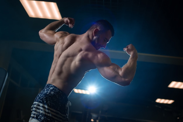 Homme sportif avec un corps musclé pose dans la salle de gym, exhibant son biceps et son dos