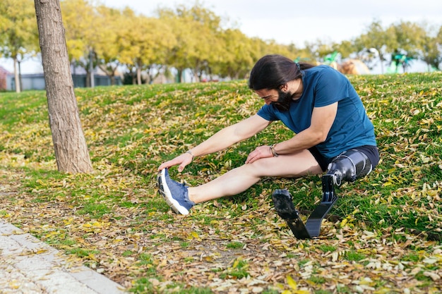 Homme sportif avec une jambe prothétique atteignant son pied tout en faisant des étirements assis dans un parc