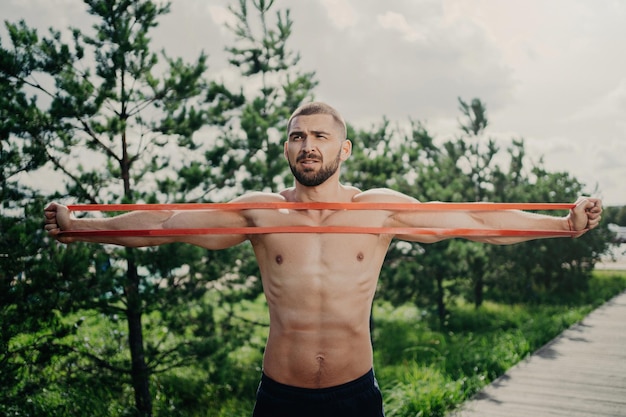 L'homme sportif avec la poitrine nue s'étire l'extenseur élastique construit des muscles forts pose à l'extérieur