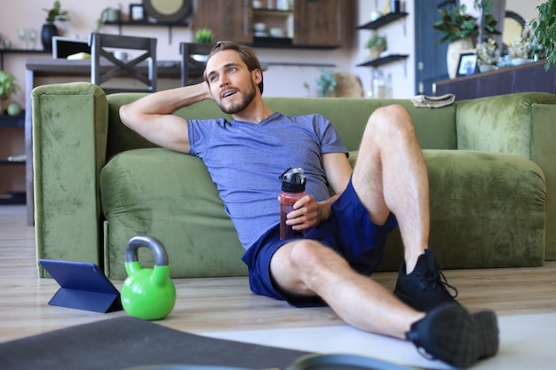 Homme sportif se reposant, ayant bu de l'eau après avoir fait de l'exercice assis sur le sol dans le salon. Fintess à la maison pendant l'auto-isolement.