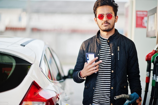 Homme sud-asiatique ou homme indien faisant le plein de sa voiture blanche à la station-service avec un téléphone portable à portée de main.