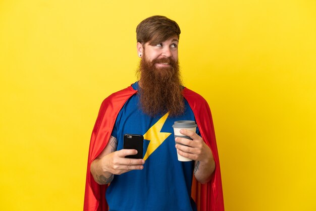 Homme super héros roux isolé sur fond jaune tenant du café à emporter et un mobile tout en pensant à quelque chose