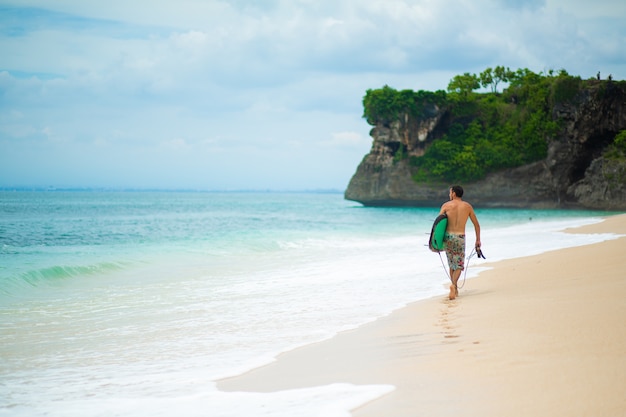 Homme de surf avec planche de surf marchant sur la plage tropicale de sable. Mode de vie sain, activités nautiques