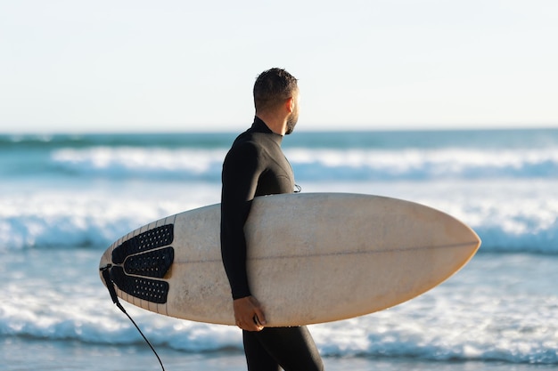 Un homme surfeur dans une combinaison de plongée marchant sur le rivage tenant une planche de surf et regardant la mer