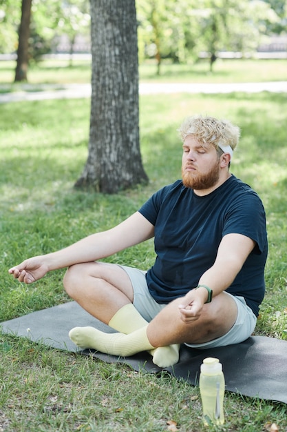 Homme en surpoids assis sur l'herbe avec les yeux fermés et se concentrant sur des exercices de yoga dans le parc