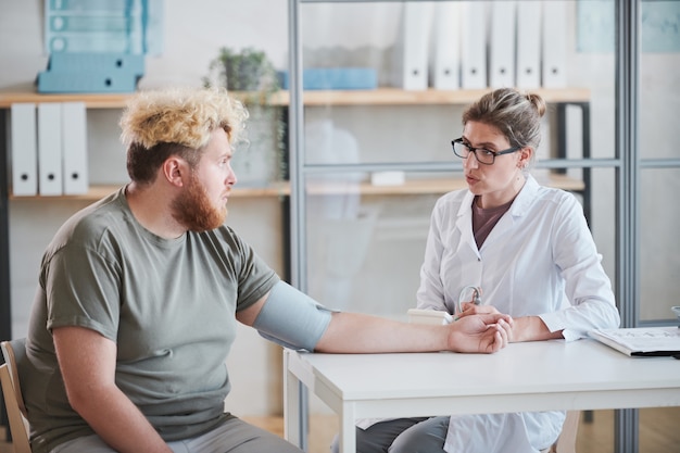 Homme en surpoids assis à table et parlant au médecin pendant qu'elle mesure sa pression pendant...