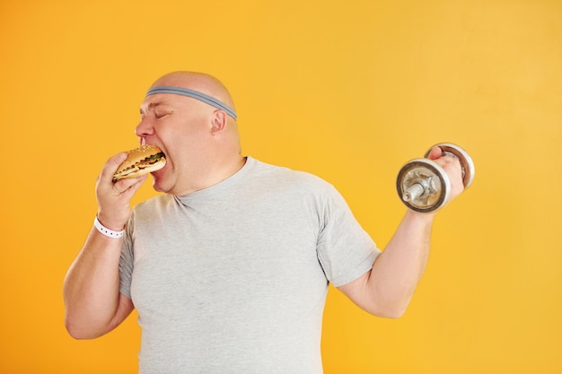 Un homme en surpoids drôle en cravate de tête sportive est titulaire d'un hamburger et d'une haltère sur fond jaune