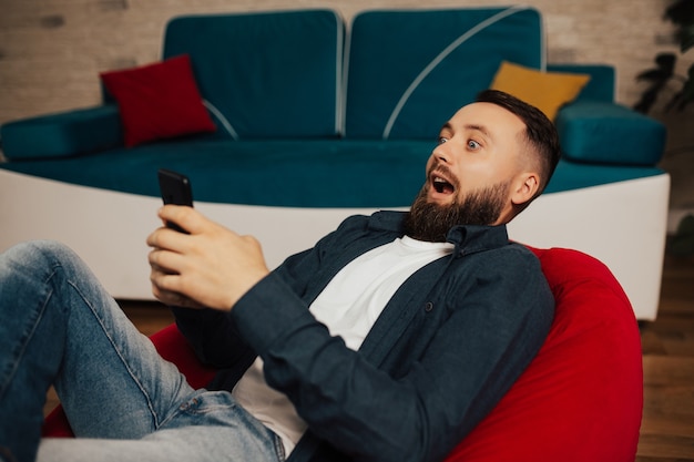 Un homme surpris, assis sur un fauteuil et tenant le smartphone, regarde l'écran de l'appareil se sent confus et choqué.