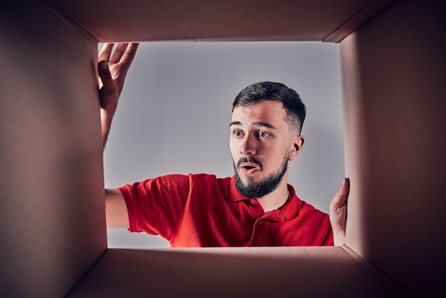 L'homme surpris déballant, ouvrant la boîte en carton et regardant à l'intérieur. Le paquet, la livraison, la surprise, le concept de mode de vie cadeau. Concepts d'émotions humaines et d'expressions faciales