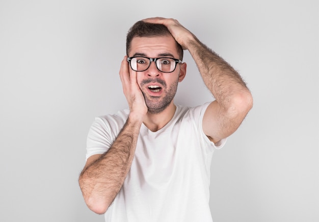 Homme surpris avec des lunettes dans un t-shirt blanc tient sa tête avec ses mains sur un mur gris