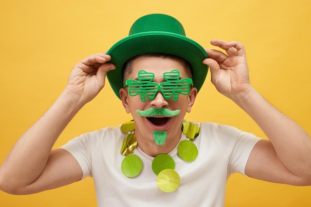 Photo un homme surpris portant un chapeau de leprechaun vert et des lunettes de trèfle pose isolé sur un fond jaune en regardant la caméra avec une expression étonnée en disant wow