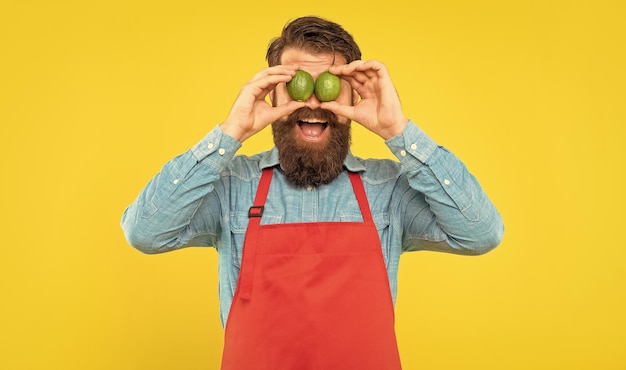 Homme surpris en tablier tenant des citrons verts frais sur les yeux vendeur de fruits fond jaune