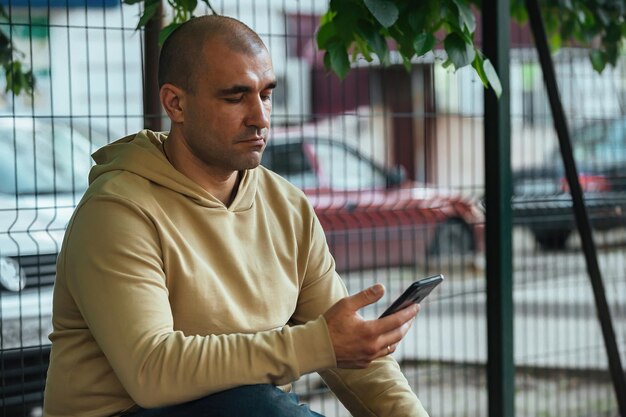 un homme en sweat-shirt léger est assis sur un banc de parc et regarde son téléphone